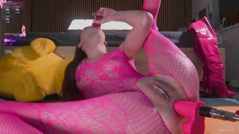 A video of a nude woman in a pink fishnet bodysuit, lying on a bed with a yellow teddy bear, and pink boots.
