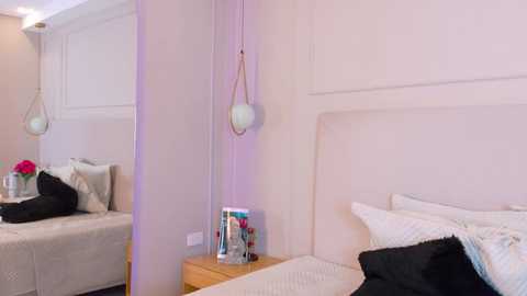 Video of a minimalist, modern bedroom with a white bed, beige walls, a wooden nightstand holding a glass vase, and two hanging pendant lights.