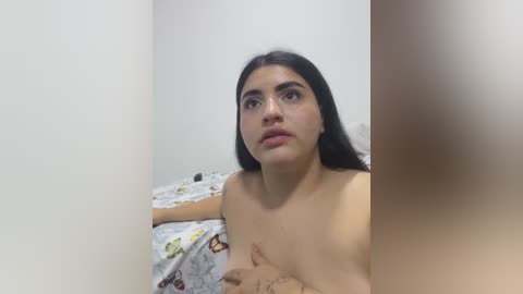 A video of a naked woman with long dark hair and medium skin tone, seated on a bed with a patterned floral sheet, looking up. Her breasts are partially obscured by her hands.