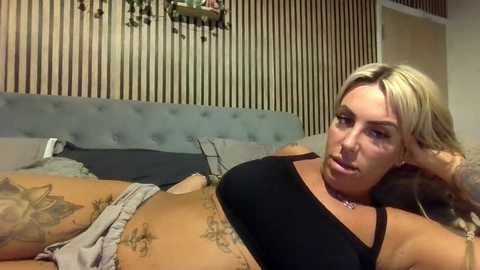 Video of a tattooed, blonde woman with a pierced nose, lying on a bed in a bedroom with a grey upholstered headboard, wearing a black tank top.