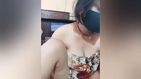 A video of a woman with light skin and medium breasts, wearing a floral dress and a black mask, sitting indoors with a TV in the background.