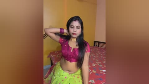 Video of a young South Asian woman with medium skin tone, wearing a bright pink short-sleeved crop top and yellow sari with floral patterns, sitting on a bed with red bedspread, in a yellow-painted room.