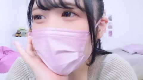A close-up video of an Asian woman with fair skin, wearing a pink face mask and a beige sweater, in a brightly lit, minimalistic room with white walls and a pink bedspread.