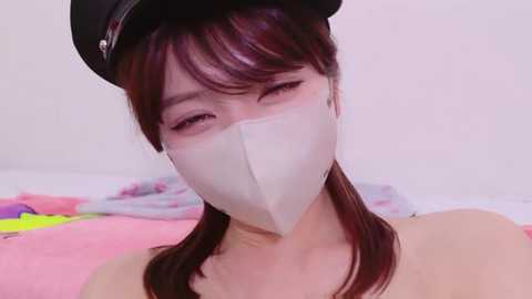 Video of a young Asian woman with long brown hair and light skin, wearing a black hat and a white face mask, lying on a colorful bedspread.