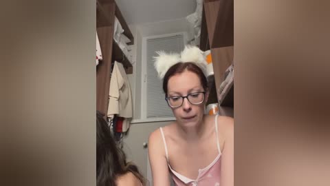 Video of a woman with glasses and a white cat ear headband, wearing a pink camisole, in a messy closet with clothes and boxes.