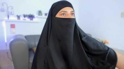 A video of a woman with light skin and brown hair, wearing a black niqab and sitting on a gray couch, in a softly lit, minimalist room.