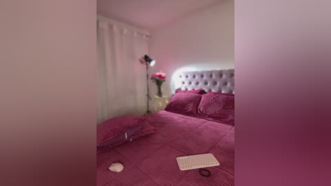 Video of a cozy bedroom with a plush, tufted purple headboard, matching bedspread, and a small white nightstand with flowers and a lamp. A laptop rests on the bed.