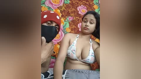 Video of a woman in a floral bikini, sitting against a backdrop of large, colorful flowers, wearing a red hat with white polka dots and a black face mask.