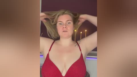 Video of a fair-skinned woman with blonde hair, wearing a red bra, standing indoors with purple walls, adjusting her hair.