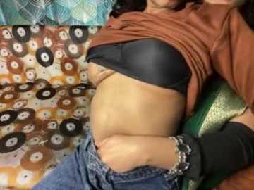 Video of a person with medium-dark skin, wearing a black bra, brown shirt, and blue jeans, reclining on a patterned couch with teal, brown, and white circles.