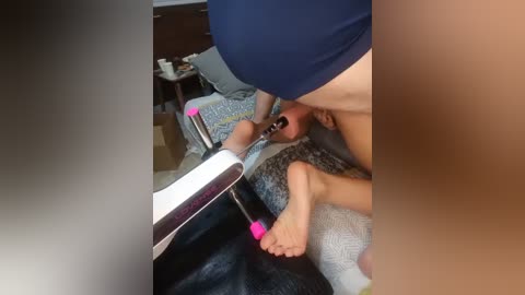 Video of a man, wearing a navy-blue cap and blue shirt, using a handheld vacuum to clean a baby's feet, lying on a grey-patterned blanket in a cluttered room with dark wooden furniture.