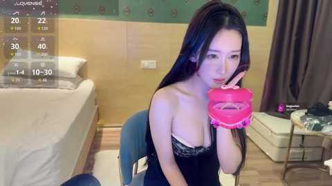 Media: A video of an Asian woman with long black hair, wearing a black lace bra, eating a heart-shaped lollipop. She sits on a blue chair in a modern, wooden-floored bedroom with a bed and green wallpaper.