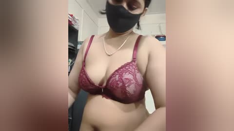 Media: Video of a woman in a maroon lace bra, black face mask, and pearl necklace, taken in a dimly lit room with a white tiled background.