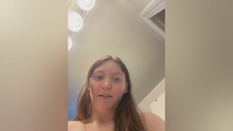 Video of a young woman with long brown hair, wearing a light-colored top, standing in a dimly lit room with white walls and a mirror with circular lights.