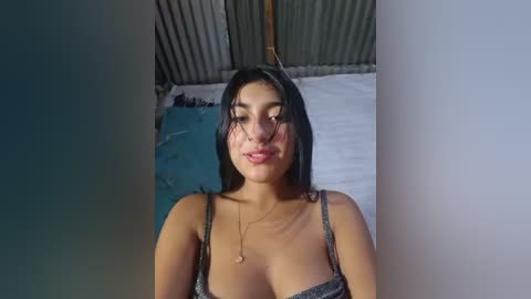 Video of a young woman with medium skin tone, dark hair, and medium breasts, wearing a sparkly spaghetti-strap top, standing in a dimly lit room with a bed and corrugated metal wall in the background.