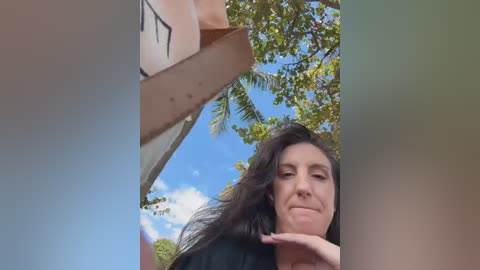 Video of a woman with long dark hair and a serene expression, standing in a bright, sunny outdoor setting with blue sky, green trees, and a beige wall in the background.