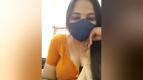 A video of an Indian woman with medium skin tone, wearing a blue mask, orange blouse, and black scarf, leaning on a table, in a dimly lit room.
