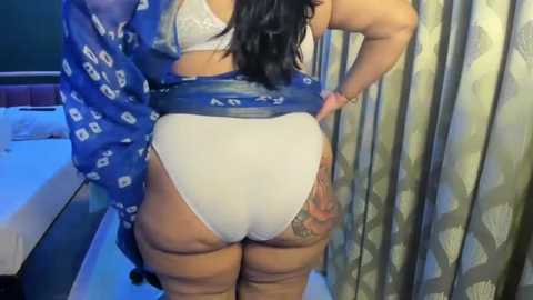 Video of a plus-sized woman with medium skin tone, black hair, and a tattoo on her right thigh, wearing white panties and a blue robe, standing in a bedroom with a bed, curtains, and a nightstand.