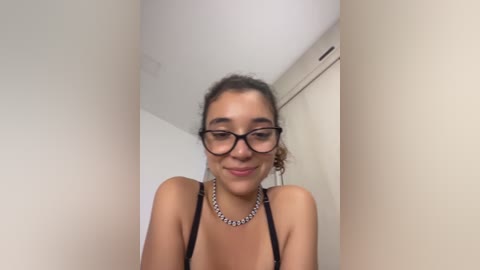 Video of a young woman with light brown skin, curly brown hair, and glasses, wearing a black bra and silver necklace. She's smiling softly in a minimalist, white-walled room.