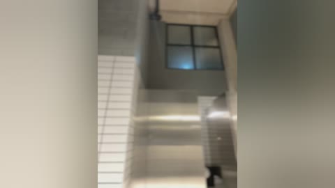 Video of a dimly lit, narrow hallway with a metallic staircase to the left, beige walls, and a small window with frosted glass at the top, creating a slightly blurred, eerie atmosphere.
