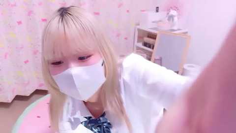 Video of a young Asian woman with long blonde hair, wearing a white face mask, white top, and blue bow tie, in a pastel-themed room with a pink floral curtain, shelves, and a wooden table.