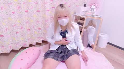 Video of a young woman with light skin, blonde hair, wearing a white face mask, white cardigan, plaid skirt, sitting on a pink bed with a watermelon pillow. Background includes a white curtain with pink patterns, wooden floor, and a bookshelf.