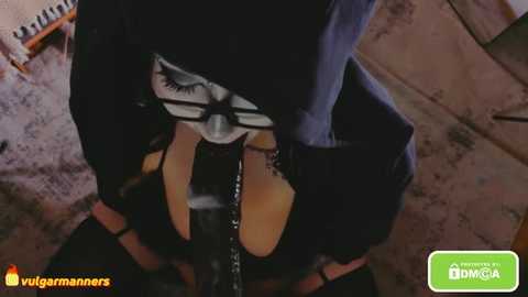 A CGI image shows a close-up of a person in a dark, tight-fitting outfit with a mask featuring a large eye, holding a whip. The background is a dimly lit room with a wooden floor.