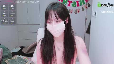 A video of a nude, fair-skinned Asian woman with long black hair and a surgical mask, sitting in a cluttered bedroom with a calendar and a \"MOM\" banner in the background.