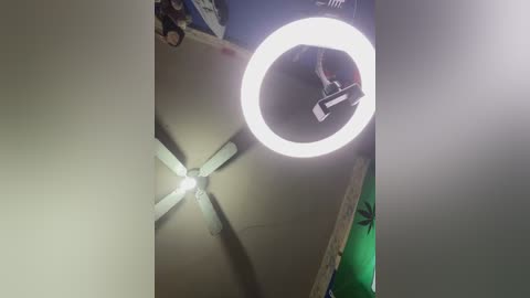 A video of a dimly lit room with a bright circular spotlight illuminating a ceiling fan, casting harsh shadows. The room appears cluttered with miscellaneous objects on a green surface in the background.