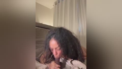 A video captures a woman with long, curly black hair, partially naked, performing oral sex on a man. The scene is dimly lit, with beige curtains and a gray bedspread in the background.
