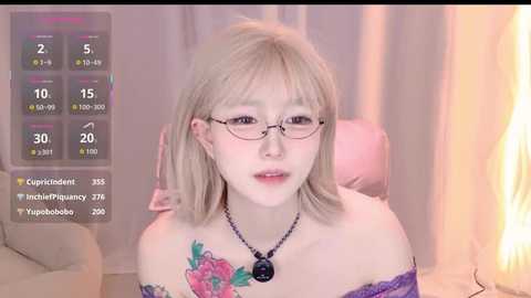 A video of a fair-skinned, petite woman with shoulder-length blonde hair, wearing glasses, a black necklace, and a purple lace off-shoulder top. She has a colorful tattoo on her left shoulder. The background shows a dimly lit room with a TV displaying a pregnancy tracker.