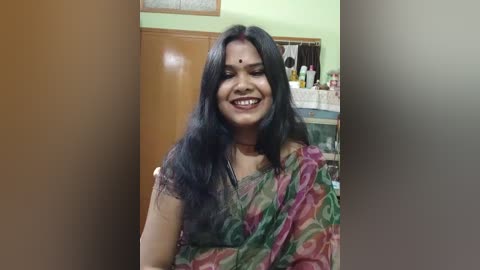 Media: Video of a South Asian woman with long black hair, wearing a colorful sari, smiling in a home setting with light green walls and a wooden door.