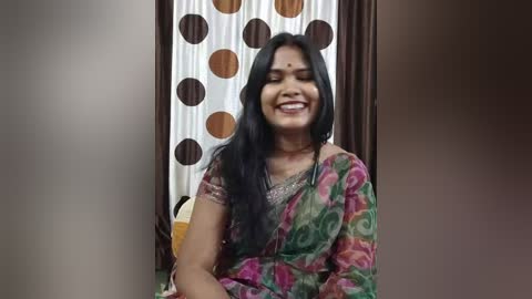 Media: Video of a joyful South Asian woman with long black hair, wearing a colorful sari, smiling widely, in a room with a polka-dotted curtain.