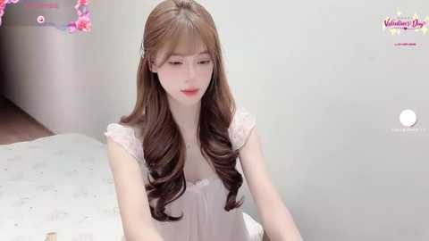 Media: Video of a young East Asian woman with long, wavy brown hair, fair skin, and pink lips, wearing a white lace dress, seated in a softly lit room with pink flowers in the background.