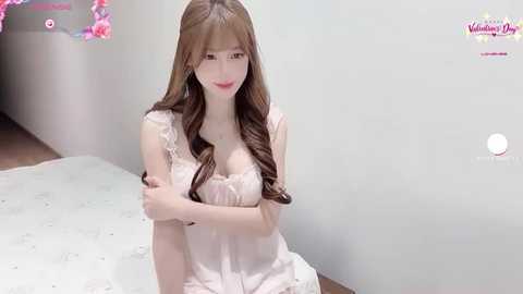 Media: A video of a young Asian woman with long brown hair, fair skin, and wearing a white lace dress, sitting on a white carpet, in a minimalistic room with a pink floral decoration in the background.