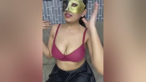 Media: A video of a woman with medium skin tone, wearing a gold masquerade mask, red bra, and black skirt, posing indoors against a tiled wall.