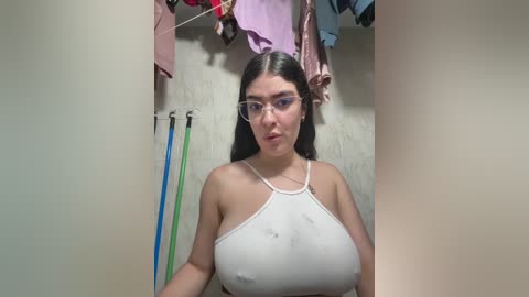 Video of a young woman with long dark hair, wearing a white spaghetti-strap top, revealing large breasts. She stands in a narrow hallway with clothes hanging on a rod, and a mop and broom leaning against the wall.