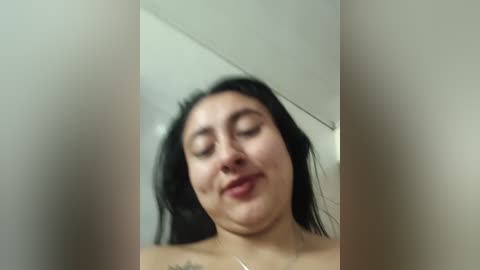 Video of a smiling, middle-aged woman with dark hair, closed eyes, and a tattoo on her upper chest, standing indoors against a plain wall.