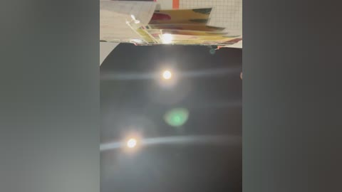 A video of a cluttered desk with multiple paper sheets, a colorful folder, and a partially visible notebook, all under harsh fluorescent lighting, creating lens flares. The background is a grid-patterned wall.