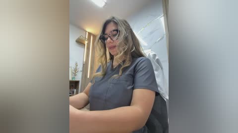 A video of a light-skinned, middle-aged woman with shoulder-length, wavy brown hair, wearing glasses and a dark blue scrub top. She sits in a dimly lit room with beige walls and a wooden door in the background.