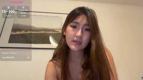 Video of an East Asian woman with long brown hair, wearing a brown top, in a modern room with a framed picture of a beach on the wall.