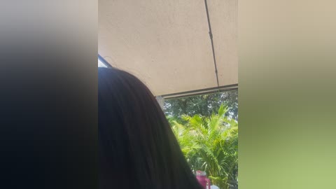 Video of a person's shoulder and back under a beige awning, with green foliage and a pink drink visible in the background.