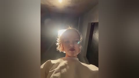 Media: Video of a young woman with short, light brown hair, wearing a beige shirt, standing in a dimly lit, narrow hallway. The image is slightly blurred, with lens flares and shadows adding an atmospheric feel.