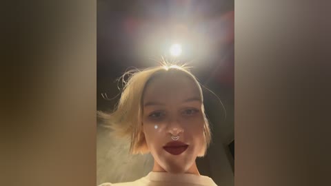 Media: Video of a young woman with short blonde hair, wearing a nose ring, and dark lipstick, taken indoors with a bright light source in the background.