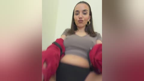 Video of a young woman with medium brown skin, straight brown hair, wearing a gray crop top, black high-waisted shorts, and a red jacket. Background is blurred, featuring a beige wall.