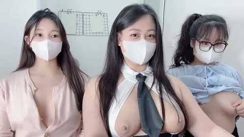 Video of three East Asian women wearing face masks, revealing their breasts in revealing outfits, with one woman wearing glasses. Background shows a white wall with a grid pattern.