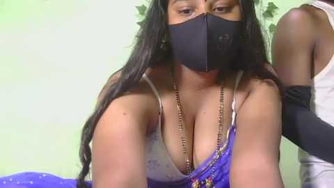 Video of a Black woman with long, straight hair, wearing a black mask, purple bra, and blue blouse, in a room with a green wall and white arm in the background.