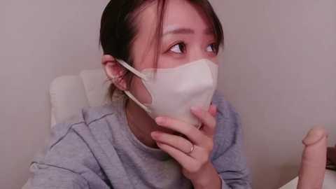 Video of an East Asian woman with dark hair, wearing a white face mask, grey sweater, and holding a large, light-colored dildo, looking concerned, in a plain, beige room.