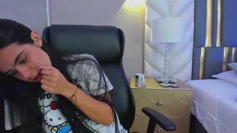 Video of a woman with long black hair, wearing a Hello Kitty T-shirt, sitting in a black office chair in a modern bedroom with a white and gold quilted headboard.