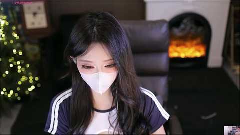 Video of an East Asian woman with long black hair, wearing a white face mask, navy Adidas shirt, and sitting near a lit Christmas tree and a fireplace.